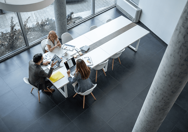 Fotografía cenital en plano picado que muestra a tres personas de negocios (dos mujeres y un hombre) sentadas en el extremo de una amplia mesa rectangular de reuniones blanca. Están rodeados de laptops, carpetas y documentos de trabajo. La oficina tiene un diseño contemporáneo con grandes ventanales, suelo de baldosas oscuras y una columna de hormigón vista.